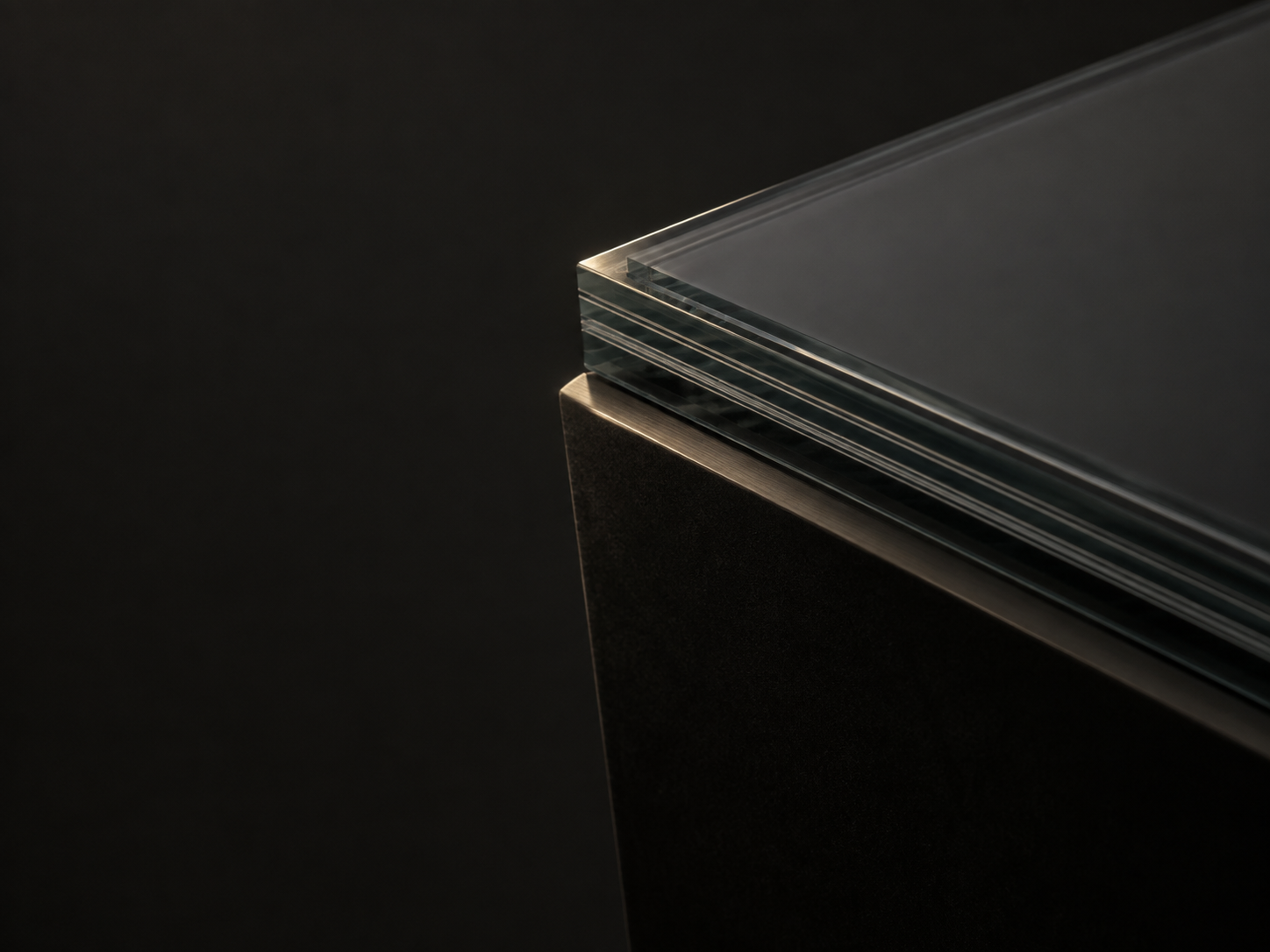 A thick slab of laminated ballistic glass resting on the corner of an opaque armor panel with a brushed steel edge